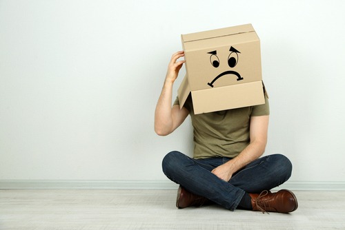 Image of a man sitting cross legged on the floor. A cardboard box with a frown face drawn on it sits on his head.