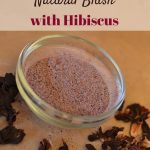 Image of a clear glass ramekin filled with blush colored powder. Dried hibiscus flowers are scattered around for decorative effect. Text overlay says, "How to Make Natural Blush with Hibiscus".