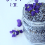 Image of a small Mason jar filled with purple colored bath salts. Top and to the sides are garnished with fresh violet flowers. Text overlay says, "Violet Sea Salt Bath Soak Recipe".