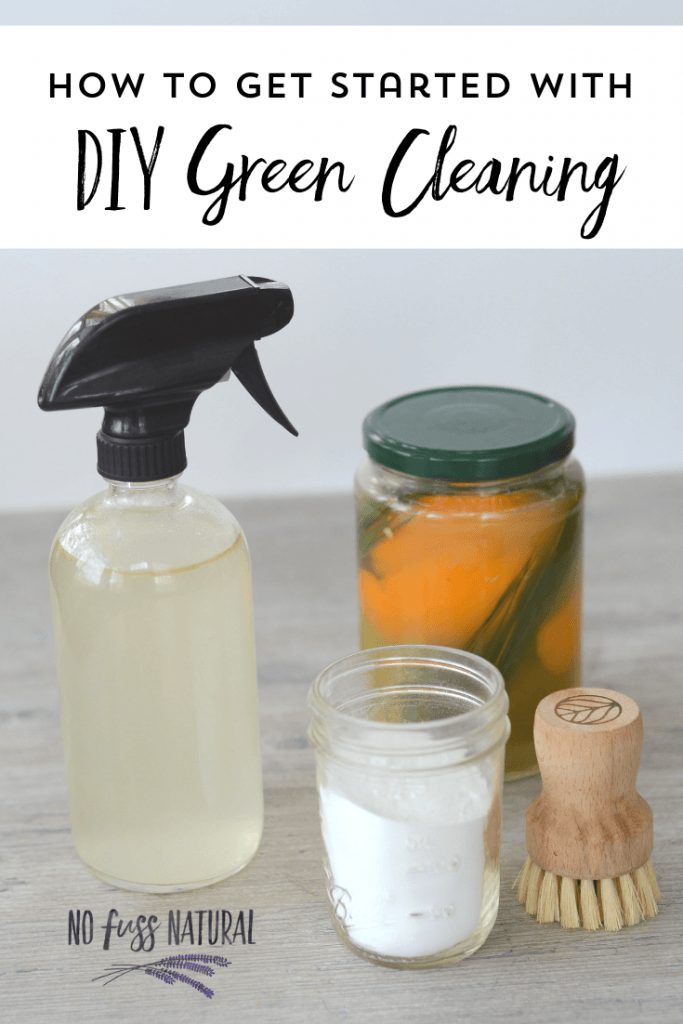 collection of green cleaning supplies: glass spray bottle with homemade cleaner, fruit peel vinegar in jar, baking soda in jar, and wooden scrubbing brush